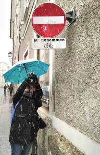 Eine Straße in Salzburg bei Schneefall. Eine Person in Winterkleidung fotografiert ein Schild auf der Straße. Eine zweite Person hinter ihr hält ihr einen Regenschirm.